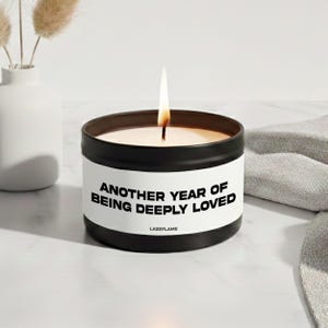 Può includere: Una candela in un barattolo di metallo nero con un'etichetta bianca che riporta il testo nero "ANOTHER YEAR OF BEING DEEPLY LOVED" e "LAZEFLAME". La candela è accesa, mostrando una fiamma singola che si alza dal centro della cera nel contenitore.