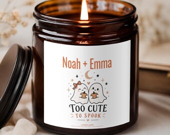 May include: A brown glass jar candle with a lit wick and a white label. The label features the text Noah + Emma, a graphic of two ghosts holding pumpkins, and the text TOO CUTE TO SPOOK. The candle has a black lid resting beside it.