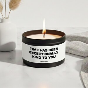 Könnte beinhalten: Eine schwarze Metalldose mit einer Kerze und einem weißen Etikett, das den schwarzen Text "TIME HAS BEEN EXCEPTIONALLY KIND TO YOU" und "LAZEFLAME" trägt. Die Kerze brennt und zeigt eine einzelne Flamme, die aus dem weißen Wachs aufsteigt.