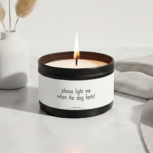 Könnte beinhalten: Eine schwarze Metalldose mit einer weißen Banderole, auf der in schwarzer Schrift "please light me when the dog farts!" und der Markenname "LAZEFLAME" steht. Die Kerze brennt mit einer sichtbaren Flamme und das Wachs ist cremefarben.