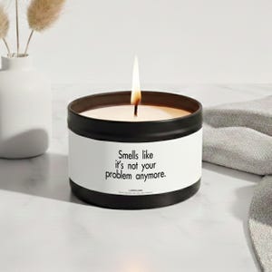 Könnte beinhalten: Eine schwarze Metalldose mit einer Kerze und einem weißen Etikett, das den schwarzen Text "Smells like it's not your problem anymore." sowie den Markennamen "LAZEFLAME" trägt. Die Kerze hat einen brennenden Docht mit sichtbarer Flamme.