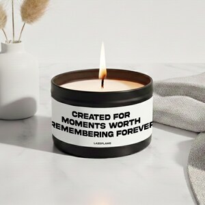 Può includere: Una candela in barattolo di metallo nero con un'etichetta bianca che riporta il testo nero CREATED FOR MOMENTS WORTH REMEMBERING FOREVER e LAZEFLAME. La candela ha uno stoppino acceso con una fiamma visibile che emette una luce calda dalla cera.