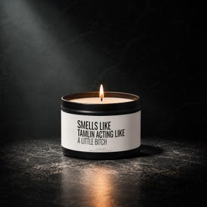 Könnte beinhalten: Eine schwarze Metalldose mit einer weißen Banderole, auf der in schwarzer Schrift steht: SMELLS LIKE TAMLIN ACTING LIKE A LITTLE BITCH. Die Kerze hat einen einzelnen brennenden Docht mit einer warmen Flamme und das Wachs ist weiß.