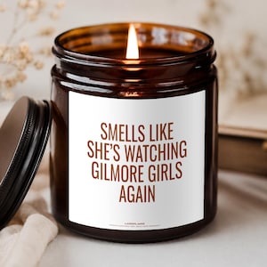 Könnte beinhalten: Eine braune Glaskerze mit einem weißen Etikett, das den braunen Text trägt: SMELLS LIKE SHE'S WATCHING GILMORE GIRLS AGAIN. Die Kerze brennt mit einer sichtbaren Flamme und der schwarze Metalldeckel liegt neben dem Glas auf einer Oberfläche.
