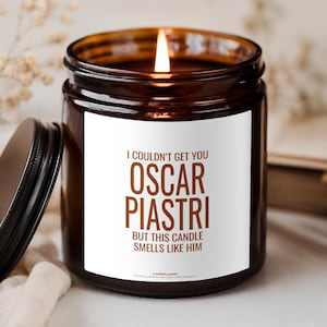 Könnte beinhalten: Eine brennende Kerze in einem dunkel bernsteinfarbenen Glasgefäß mit schwarzem Deckel. Das weiße Etikett trägt den braunen Text: I COULDN'T GET YOU OSCAR PIASTRI BUT THIS CANDLE SMELLS LIKE HIM. Die Kerze hat eine sichtbare Flamme in einem zylindrischen Gefäß.