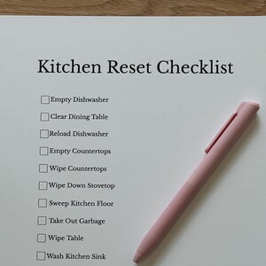 Peut inclure: Une feuille de papier blanche avec une "Kitchen Reset Checklist" imprimée en noir. La liste comprend des tâches telles que vider le lave-vaisselle et essuyer les comptoirs. Un stylo rose est posé sur le papier. La phrase "Progress Over Perfection" est en bas.
