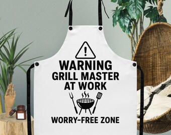 Puede incluir: Delantal blanco con correas negras y el texto "WARNING GRILL MASTER AT WORK WORRY-FREE ZONE". El delantal presenta un símbolo de advertencia, una parrilla, un tenedor y una espátula.
