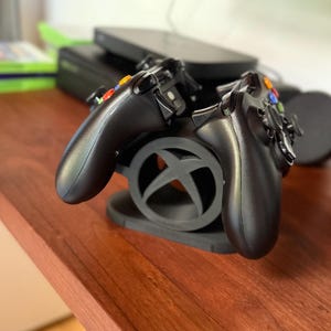 Puede incluir: Un soporte negro para mandos de Xbox que sostiene dos mandos negros. El soporte presenta el logotipo de Xbox. Los mandos tienen botones de colores y almohadillas direccionales. El soporte y los mandos están sobre una superficie de madera.