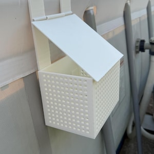 May include: A white, rectangular pool accessory with a hinged lid and a perforated basket design. The item is designed to hang on the side of a pool. The background shows a pool and surrounding environment.