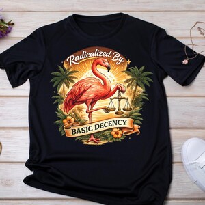 May include: Black t-shirt with a flamingo design holding scales, and the text "Radicalized By Basic Decency." The image includes palm trees, a sunset, and tropical flowers. The flamingo is rendered in pink and orange hues.