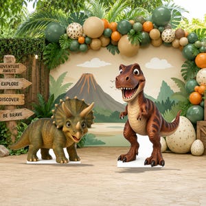 May include: Two dinosaur standees featuring a brown Tyrannosaurus Rex and a green Triceratops. The background displays a volcano scene with a balloon arch and a wooden signpost with the text Adventure Awaits, Explore, Discover, Be Brave.
