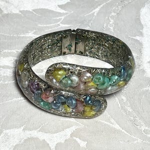 May include: A bypass-style cuff bracelet featuring a silver-toned textured metal base embedded with a collection of small, iridescent, multi-colored seashells in shades of pink, yellow, blue, and green, secured with a hinged metal clasp.