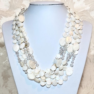 May include: A multi-strand white necklace with a layered design. The necklace features large, flat, round, and oval-shaped beads, interspersed with clear crystal accents. The necklace is displayed on a white mannequin bust.