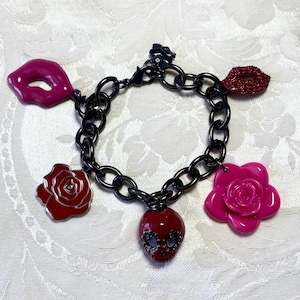 May include: A black chain link charm bracelet with various charms. Charms include a red rose, a pink rose, a red skull, and two sets of lips in pink and red. The bracelet is on a white surface.