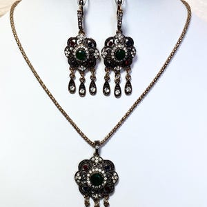 May include: A vintage-style jewelry set featuring a necklace and matching earrings. The set has a floral design with a central green stone, surrounded by smaller red, blue, and clear stones. The earrings and pendant have dangling teardrop accents.