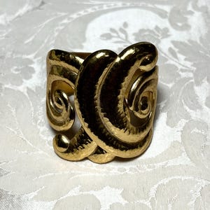 May include: A gold-toned ring featuring a wide, open-ended band with a prominent, hammered texture. The design consists of two large, swirling, spiral-shaped elements that overlap and curve around the center, creating a bold, sculptural appearance.