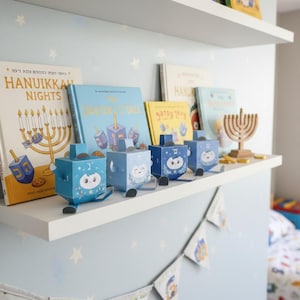 Puede incluir: Una estantería blanca exhibe libros temáticos de Janucá y dreidels decorativos. Los libros incluyen títulos como "Hanukkah Nights" y "The Dreidel's Tale". Los dreidels son azules con diseños de búhos. Una menorá de madera y guirnaldas completan la exhibición.