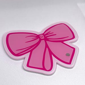 Puede incluir: Un objeto plano con forma de lazo que presenta una base de color rosa claro con un contorno de color rosa intenso y un detalle central. El artículo tiene un aspecto liso y superpuesto con un pequeño imán metálico circular en la parte inferior derecha.