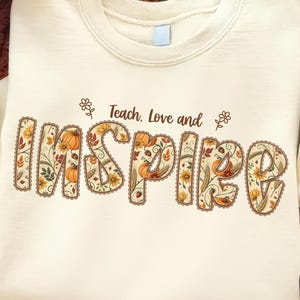 Op de afbeelding: Een crèmekleurig sweatshirt met ronde hals en de tekst "Teach, Love and INSPIRE" in het bruin. Het woord "INSPIRE" is gevuld met een patroon van oranje pompoenen, gele zonnebloemen, bruine eikels en groene bladeren met een geschulpte rand.