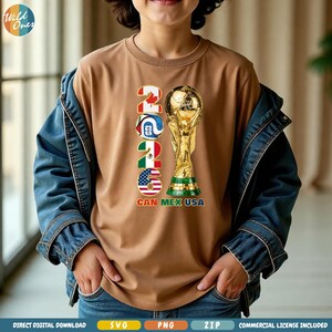 May include: A brown t-shirt featuring the text "2026" with the flags of Canada, Mexico, and the USA, along with a golden trophy. The shirt also includes the text "CAN MEX USA". The shirt is worn by a person wearing a blue denim jacket.