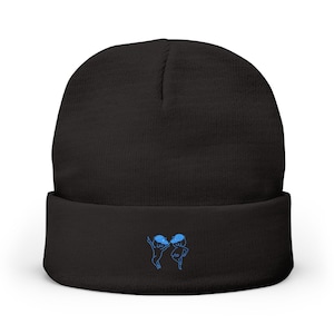 May include: Black beanie hat with a folded brim. The hat has a blue embroidered design of two dancing figures. The figures are stylized with rounded heads and bodies. The beanie is made of a soft, knitted material.