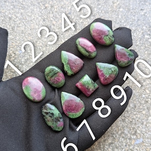 Könnte beinhalten: Zehn polierte Rubin-Zoisit-Edelsteine in verschiedenen Formen, darunter oval, tropfenförmig und rautenförmig, mit gesprenkelten Mustern in tiefem Magenta und Waldgrün. Jeder Stein ist mit einer weißen Zahl von 1 bis 10 zur Identifizierung versehen.