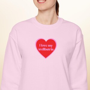 May include: A light pink sweatshirt with a red heart graphic. The heart contains the text "I love my wellbutrin" in a retro font. The sweatshirt is a crew neck style.