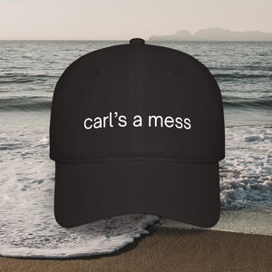 May include: Black baseball cap with the text "carl's a mess" in white. The cap is set against a beach scene with the ocean and a boat in the background.