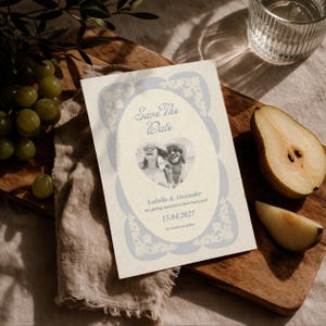 Könnte beinhalten: Eine Hochzeitsankündigungskarte mit dem Text "Save The Date" und einem Foto eines Paares. Die Karte hat einen hellblauen floralen Rand und die Namen "Isabella & Alexander" sind unter dem Foto aufgedruckt. Das Datum ist der 15. April 2027.