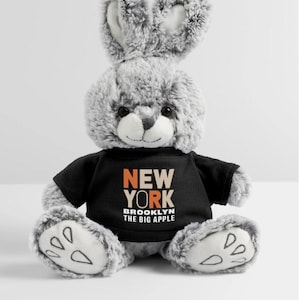 May include: A plush gray bunny wearing a black t-shirt with the words "NEW YORK BROOKLYN THE BIG APPLE" in orange and white. The bunny has large ears, a small nose, and white paw pads.