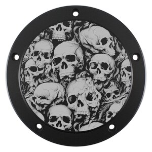 Puede incluir: Placa metálica negra y circular con un diseño detallado de numerosas calaveras en tonos grises y blancos. La placa tiene un acabado mate e incluye varios orificios de montaje espaciados.