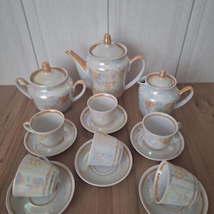 May include: A vintage tea set featuring a pearlescent finish with gold trim. The set includes a teapot, sugar bowl, creamer, and several teacups with saucers. The tea set is arranged on a wooden surface.