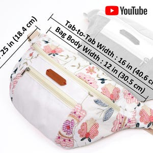 May include: White fanny pack with floral embroidery and two zippered compartments. The bag measures 7.25 inches tall and 16 inches wide.
