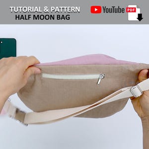 May include: A half moon shaped bag with a zippered pocket and a long strap. The bag is made of a light brown fabric with a pink lining. The bag is being held open by a hand.
