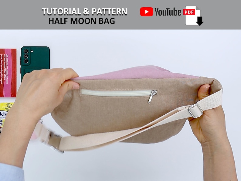 May include: A half moon shaped bag with a zippered pocket and a long strap. The bag is made of a light brown fabric with a pink lining. The bag is being held open by a hand.