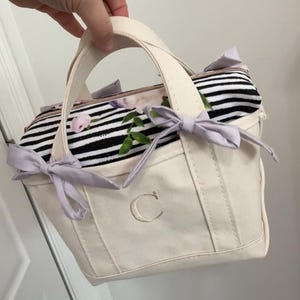 May include: Cream-colored tote bag with a floral and striped interior. The bag features a light purple bow on each side and a stitched "C" on the front. The handles are cream-colored, and the interior lining has a floral pattern with pink roses.