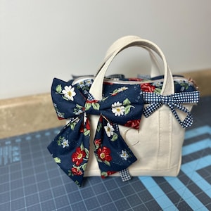 May include: A cream-colored tote bag with two handles and a floral bow. The bow is navy blue with red roses and white flowers. A smaller blue and white gingham bow is also attached. The bag is sitting on a blue cutting mat.