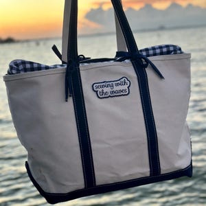 May include: Cream-colored tote bag with navy blue accents and handles. The bag features the text "sewing with the waves" on a patch. The bag is open, revealing a blue and white checkered fabric inside. The background is a sunset over the water.