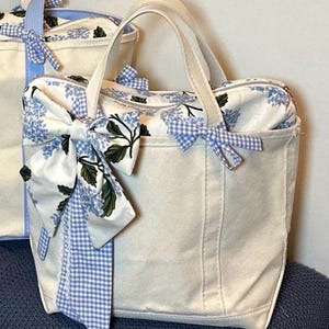 May include: A cream-colored canvas tote bag featuring a zipper closure, two top handles, and a floral pattern in blue and green. The bag is decorated with a large fabric bow made of matching floral fabric and a blue and white gingham ribbon trim.