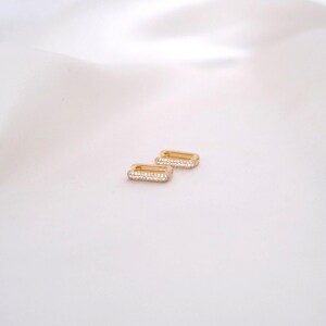 May include: A pair of gold-colored rectangular hoop earrings. Each earring is encrusted with small, clear crystals along the front edge. The earrings are displayed on a white, satin-like surface.