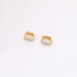May include: Two gold-colored square hoop earrings with a row of small, clear stones set along the bottom edge. The earrings are displayed on a white background, showcasing their simple yet elegant design. These earrings are a classic jewelry item.