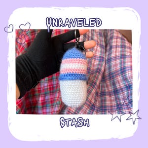 May include: A crocheted keychain in the shape of a capsule, featuring blue, pink, and white stripes. The keychain is attached to a silver ring. The word "UNRAVELED" is written at the top, and "STASH" at the bottom.