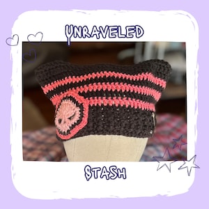 Peut inclure: Un bonnet en crochet marron foncé et rose avec des oreilles de chat. Le chapeau porte l'inscription "UNRAVELED" en haut et "STASH" en bas. Il présente des rayures roses et un écusson rose en forme de crâne.