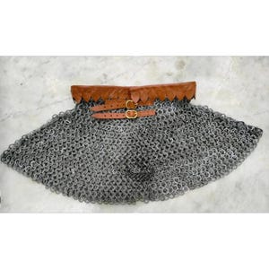 Könnte beinhalten: Ein silberner Kettenrock mit einem braunen Ledergürtel und einer Schnalle. Der Kettenrock besteht aus kleinen, ineinandergreifenden Metallringen. Der Gürtel hat zwei goldfarbene Schnallen und ist am Kettenrock befestigt.