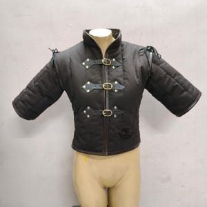 May include: Black padded medieval gambeson with three leather straps and brass buckles. The short-sleeved jacket has a high collar and is displayed on a mannequin. The jacket has a logo on the lower right side.