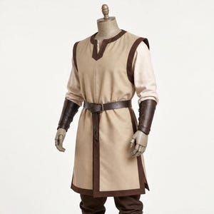 May include: A beige sleeveless tunic with dark brown trim and a V-neckline, worn over a long-sleeved beige shirt. The outfit includes a dark brown belt with a buckle and matching dark brown leather-like forearm bracers on each arm.