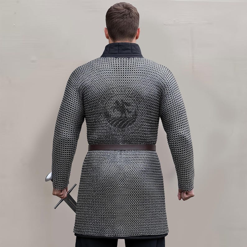 May include: A silver chainmail shirt with long sleeves and a brown leather belt. The shirt has a black collar and a design on the back. A sword is held in the right hand. This medieval garment is suitable for historical reenactments.