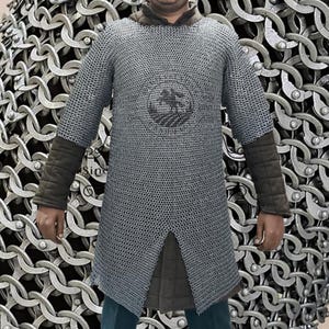 Puede incluir: Una camisa de cota de malla plateada con mangas largas y dobladillo dividido. La camisa tiene puños acolchados marrones y un cuello marrón. Un emblema circular es visible en el pecho. La camisa está hecha de pequeños anillos de metal.