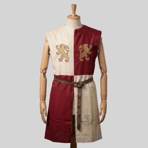 May include: A sleeveless tunic featuring a quartered design of cream and deep red fabric. Each section displays a gold embroidered lion emblem. The garment is cinched at the waist with a brown leather belt featuring a metal buckle and strap.