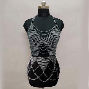 May include: A silver chainmail top and skirt set displayed on a black mannequin. The halter top features a V-neck design with layered chain details. The skirt has a chain fringe along the bottom edge. The set is made of small, interlocking metal rings.